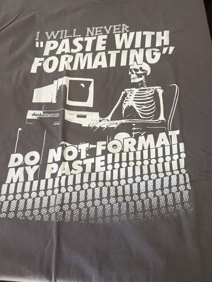 A dark t-shirt featuring a graphic of a skeleton sitting at a computer. The text reads: "I WILL NEVER 'PASTE WITH FORMATTING', DO NOT FORMAT MY PASTE!!!!!!!!!!"