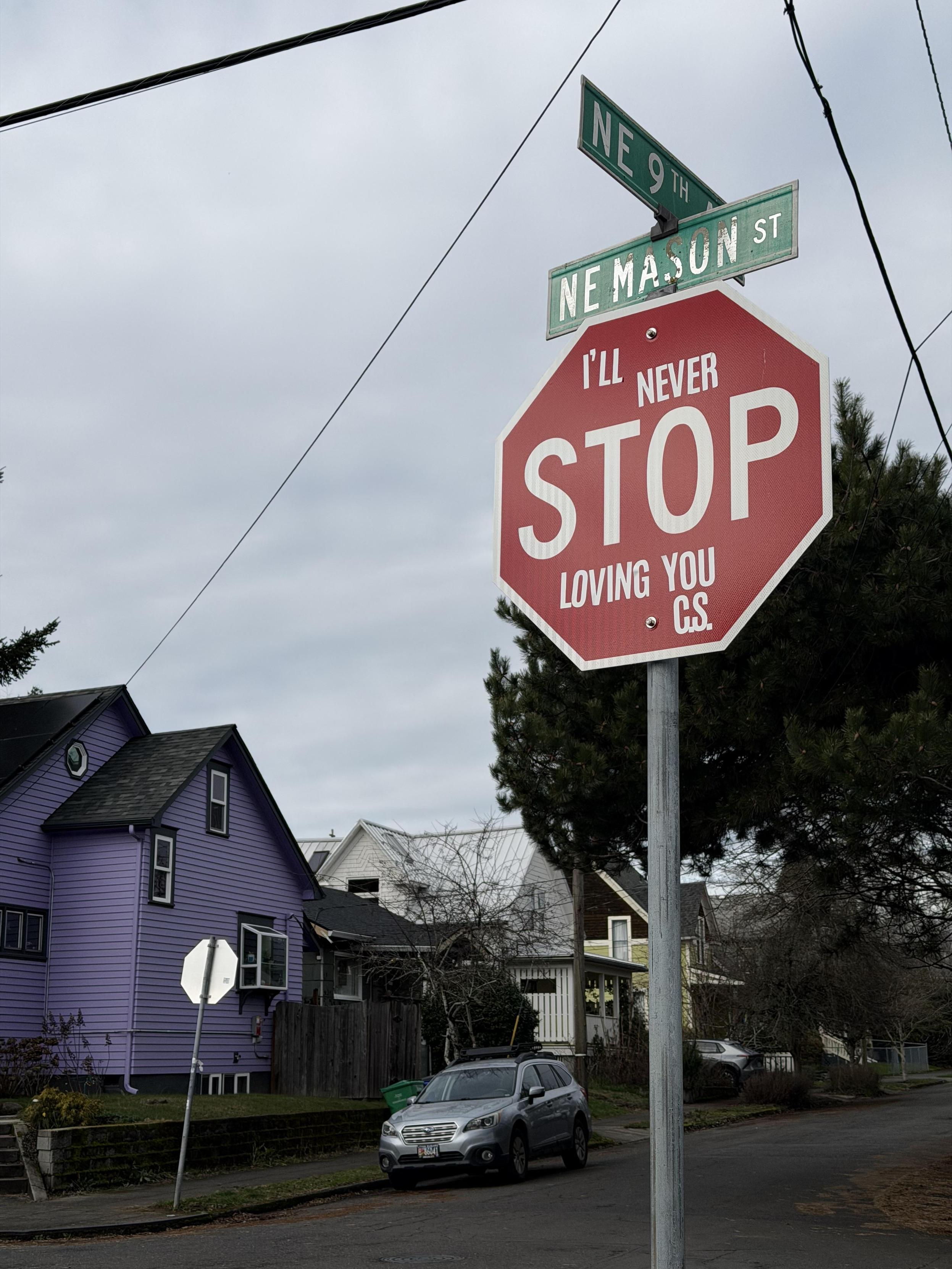 A stop sign on the corner of a street with stickers before and after STOP so that the sign reads โIโll never STOP loving you, C.S.โ