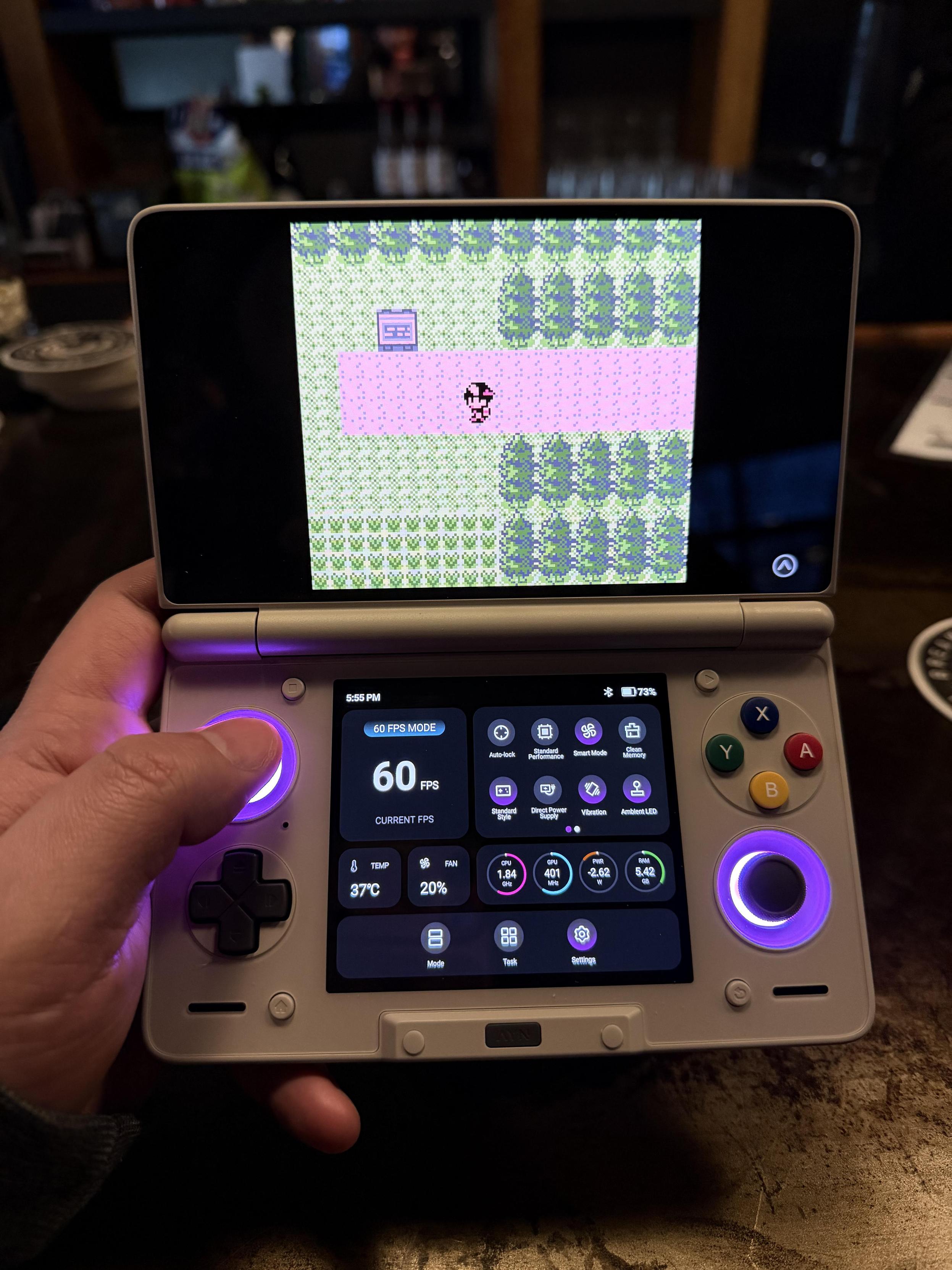 An AYN Thor (a dual-screen Android handheld) being held left-handed; the top screen shows Pokรฉmon Polished Crystal while the bottom screen shows system stats like temperature and CPU/GPU speeds