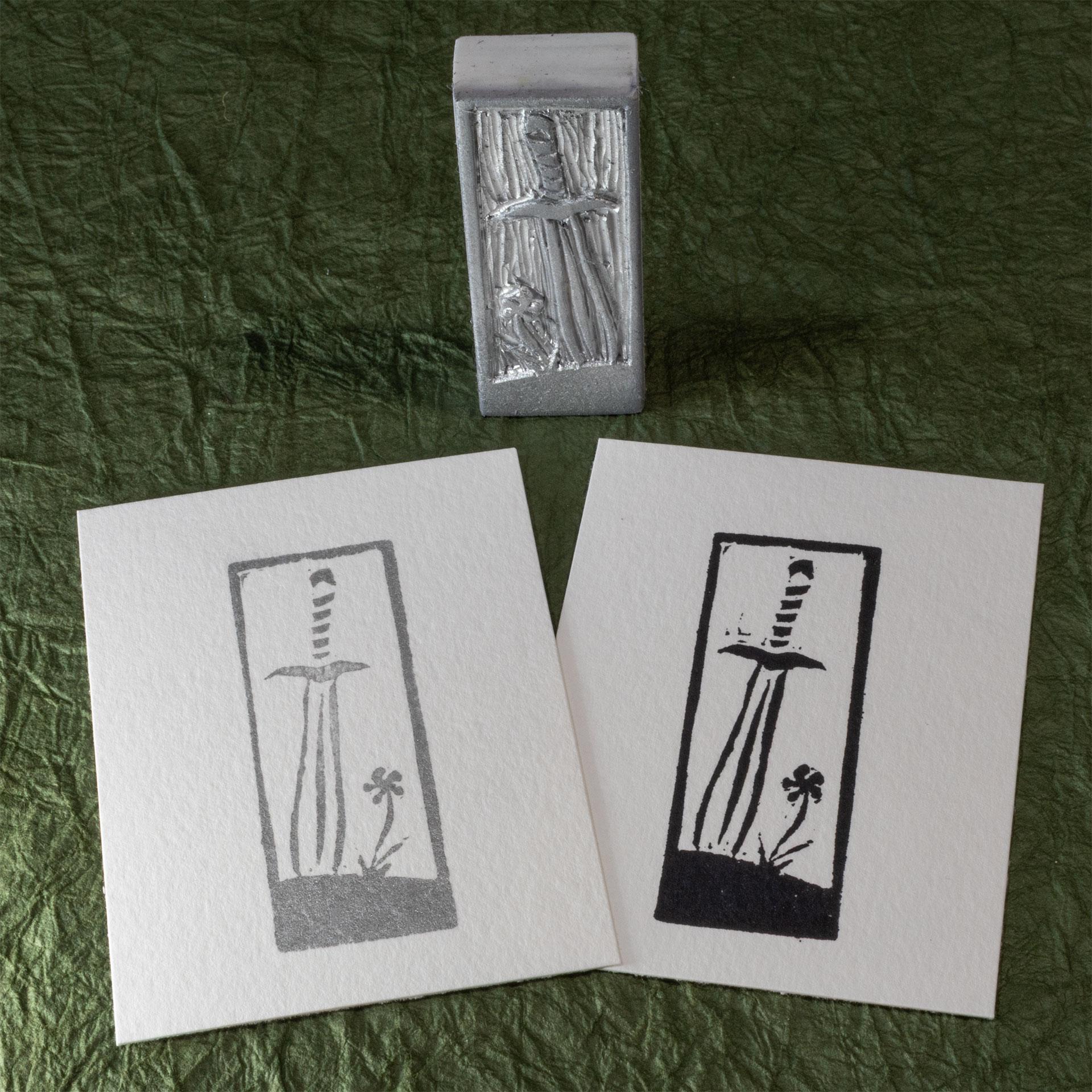 A stamp hand-carved from a blocky white eraser stands on one of the narrow sides on a background of crinkled dark green paper (momigami) behind two impressions made from it, one in black ink, one in silver ink. The design is a leaf-blade sword with the tip stuck into the ground next to a single flower.