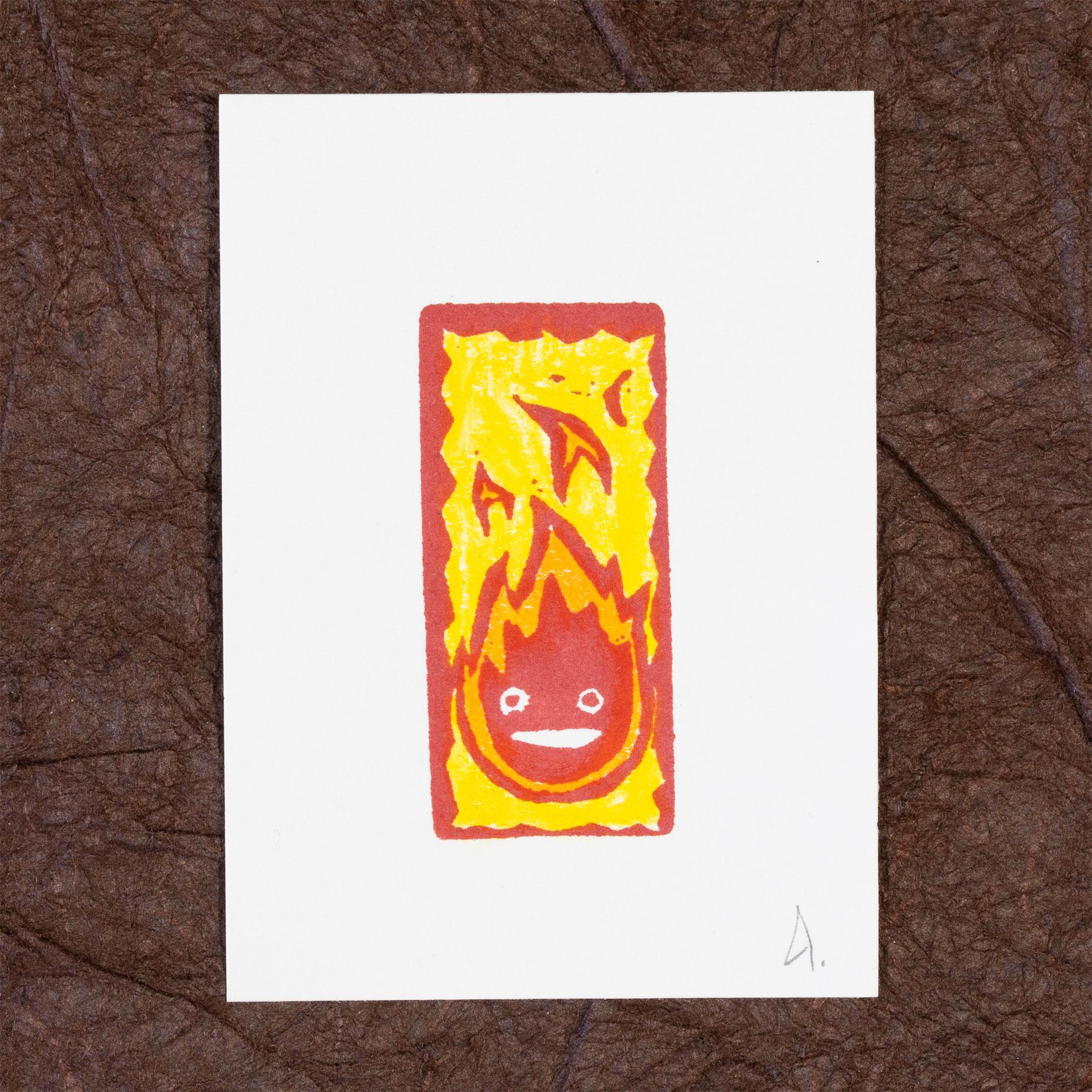 A white rectangle of paper imprinted with Calcifer, a stylized flame with two eyes and a mouth. The print has also been colored in with yellow and orange watercolor pencils. The print is signed with a small A and is resting on a background of crinkled brown paper (momigami).